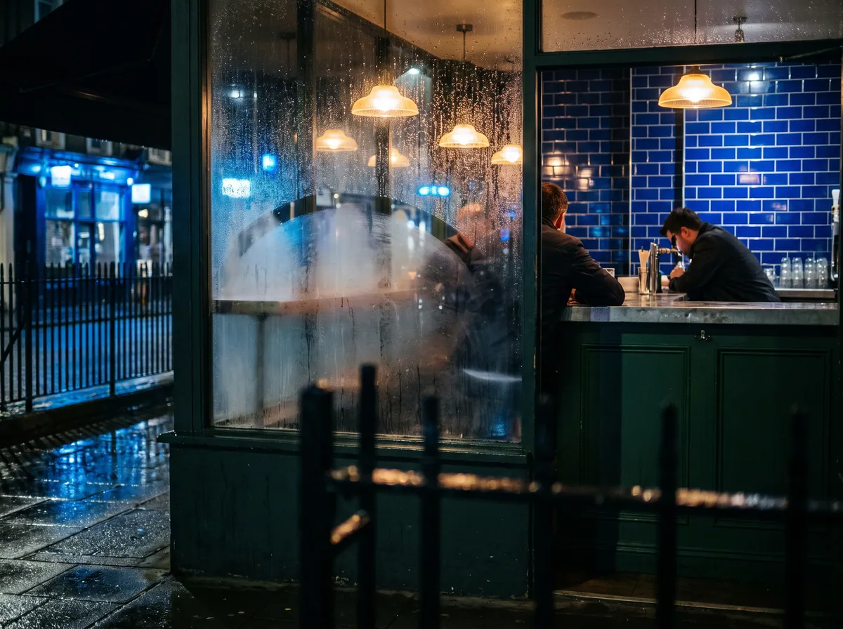 Two men at a bar counter under warm pendant lamps, seen at night through a misted window from a wet street.