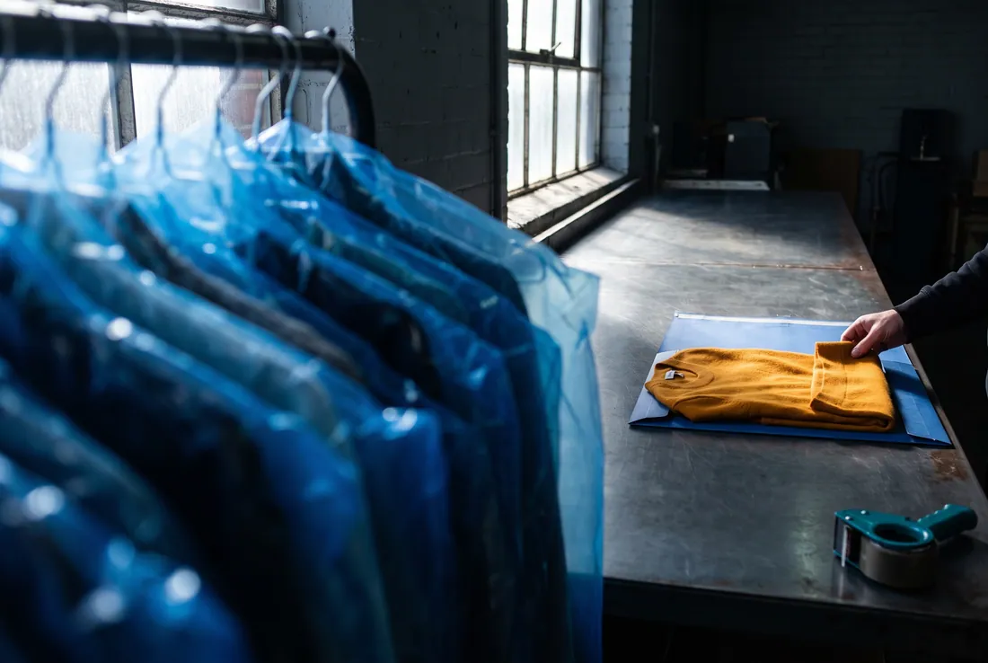 A hand smooths a folded mustard knit onto blue wrapping on a metal table, a rail of covered garments alongside.